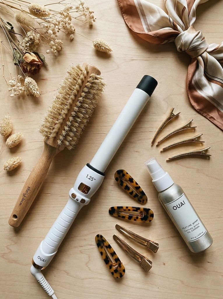 Flat lay of a round brush, curling wand, texture spray, and hair clips for styling curtain bangs