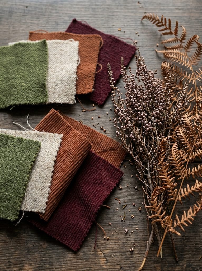 Wool and corduroy swatches in moss, oatmeal, rust, and burgundy beside dried heather and bracken