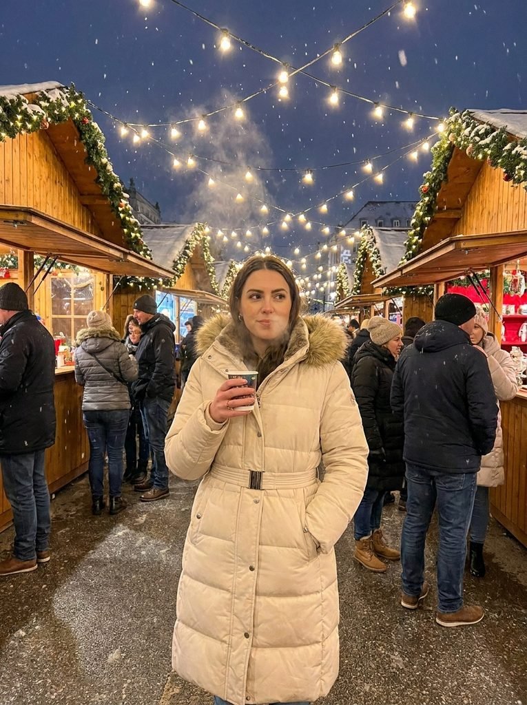 woman in cream belted puffer coat with hood standing under string lights at a freezing Christmas market