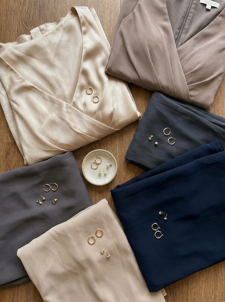 Flat lay of champagne, taupe, grey, and navy bridesmaid dresses with gold earrings