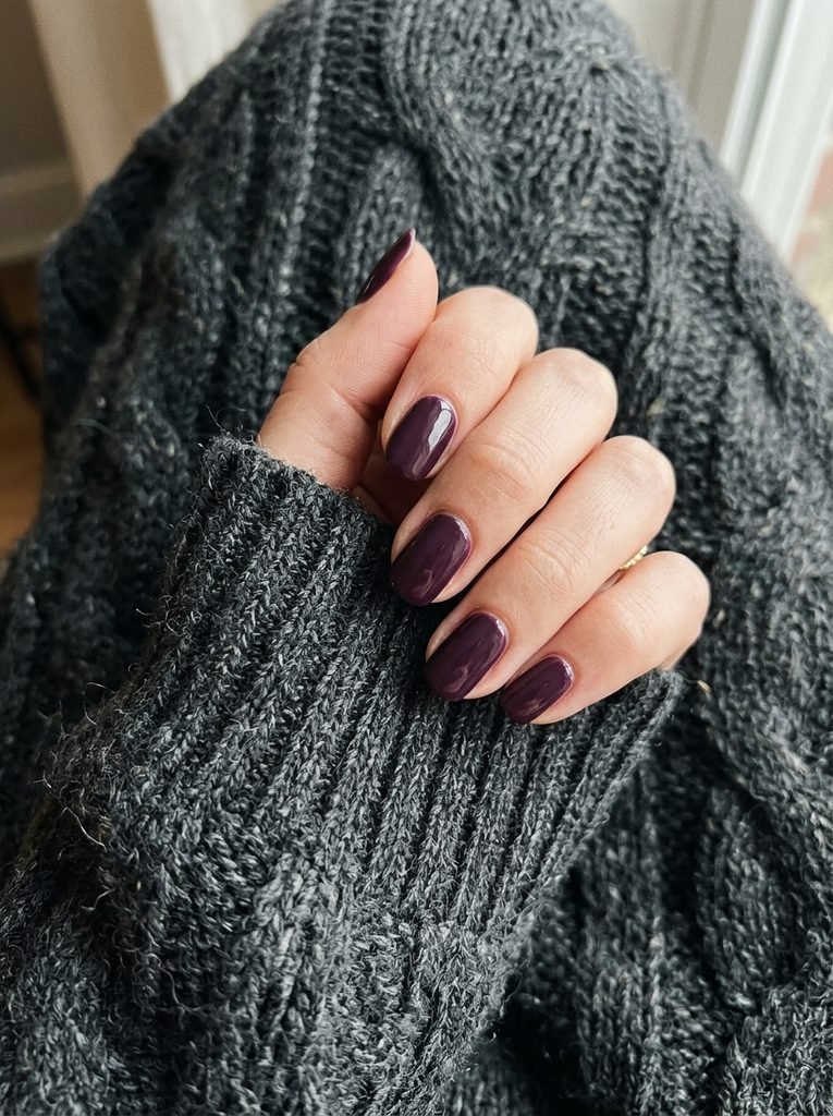 Deep plum nail polish on a hand against a charcoal grey chunky knit sweater