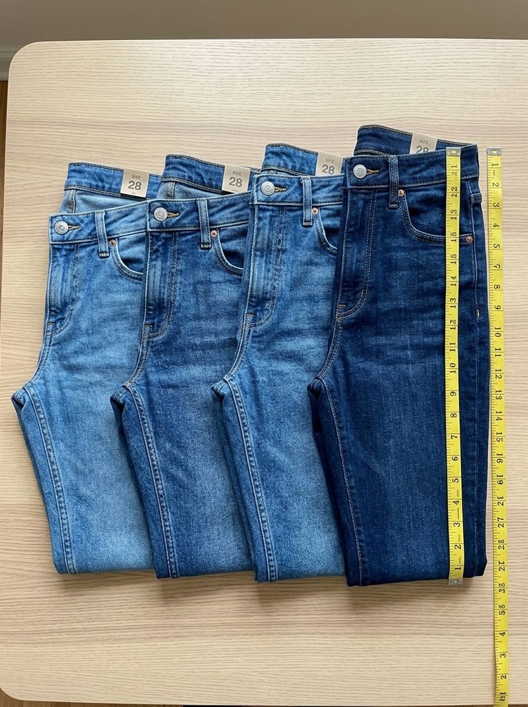 Four folded pairs of jeans in the same size showing different waistband heights, with a tape measure beside them